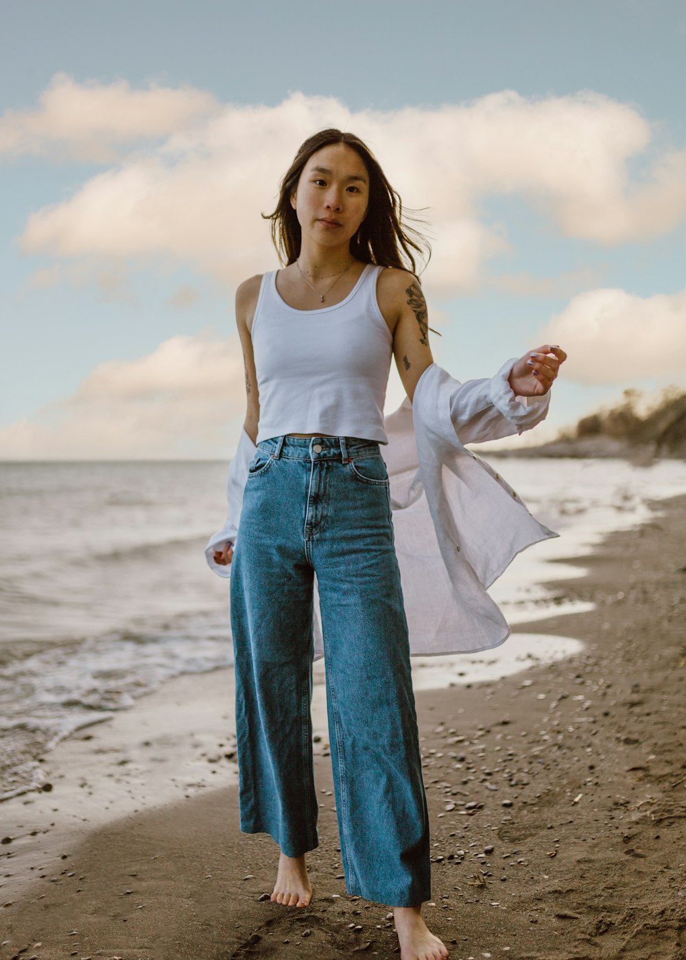 Mujer con camiseta blanca sin mangas y jeans de mezclilla azules de pie en la playa durante el día