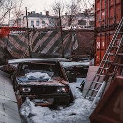 An abandoned car is partially covered in snow and surrounded by debris in an industrial or construction site. A rusted metal ladder leans against red shipping containers, while the background reveals a mix of old buildings and bare trees. The scene appears cluttered and neglected.