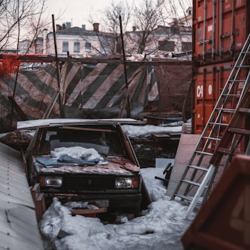 An abandoned car is partially covered in snow and surrounded by debris in an industrial or construction site. A rusted metal ladder leans against red shipping containers, while the background reveals a mix of old buildings and bare trees. The scene appears cluttered and neglected.
