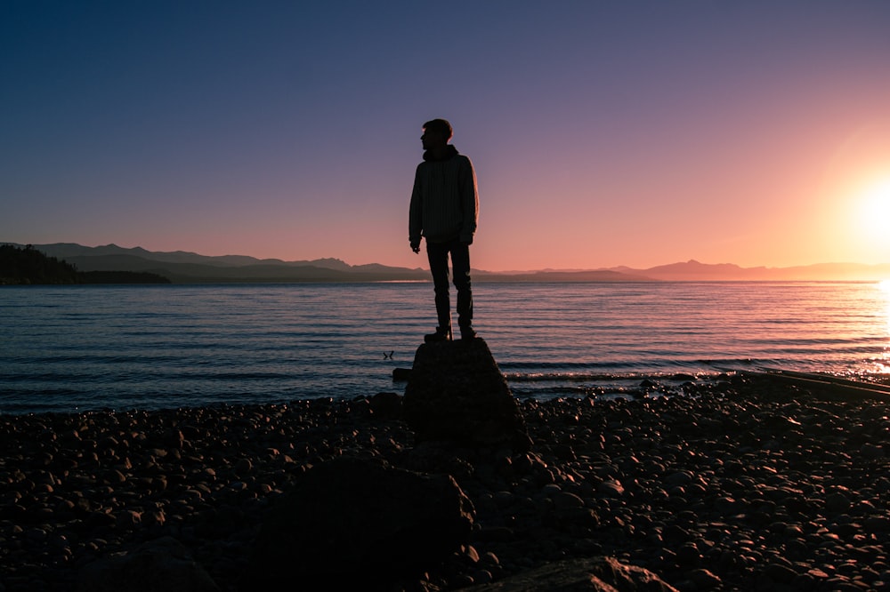 silhouette of man standing on rocky shore during sunset
