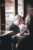 An older man with gray hair is sitting and reading a newspaper in a calm and cozy indoor space. The setting appears to be a cafe, as there are wooden furniture and large windows with outdoor patio umbrellas visible. Another person is seated nearby, slightly out of focus.
