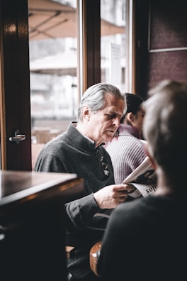 An older man with gray hair is sitting and reading a newspaper in a calm and cozy indoor space. The setting appears to be a cafe, as there are wooden furniture and large windows with outdoor patio umbrellas visible. Another person is seated nearby, slightly out of focus.