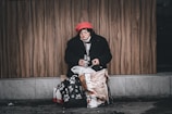 A person is sitting on a sidewalk against a wooden wall, wearing a red hat and a black coat. There are bags around them, and they appear to be holding a small bottle. The scene conveys a sense of solitude and vulnerability.