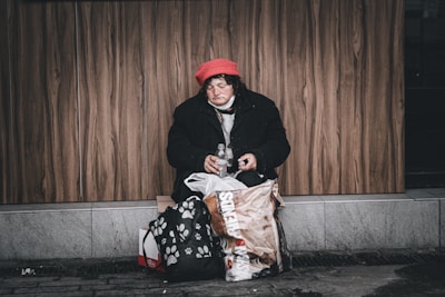 A person is sitting on a sidewalk against a wooden wall, wearing a red hat and a black coat. There are bags around them, and they appear to be holding a small bottle. The scene conveys a sense of solitude and vulnerability.