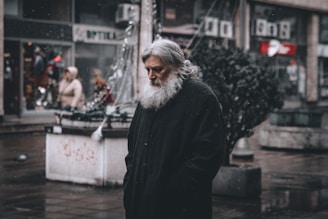 An older man with a long white beard is standing on a wet street. He wears a dark cloak-like garment. The background shows a city setting with other people walking by, some shop signs, and a few small trees. The atmosphere suggests cold weather, possibly with light snow or rain.