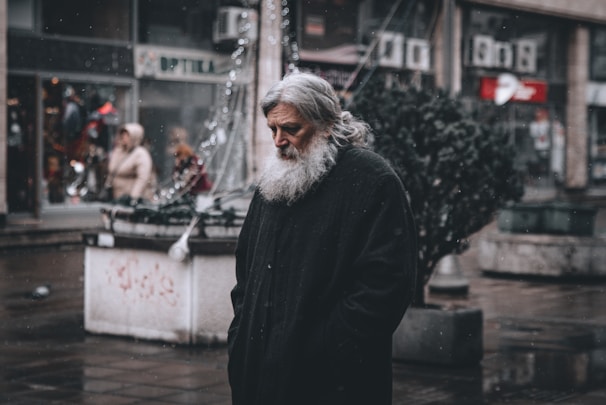 An older man with a long white beard is standing on a wet street. He wears a dark cloak-like garment. The background shows a city setting with other people walking by, some shop signs, and a few small trees. The atmosphere suggests cold weather, possibly with light snow or rain.