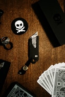 A tactical knife with intricate designs is placed on a wooden surface. Surrounding it are playing cards, a black ring, and black circular coasters featuring a white skull and crossbones symbol. The setup is complemented by a black box also adorned with the skull motif.
