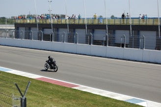 A single motorcyclist dressed in black gear rides on a racetrack with a spectator area on the right. The track is bordered by a grassy area and a series of garages with spectators observing from an elevated position behind a barrier.