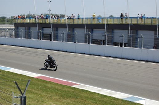 A single motorcyclist dressed in black gear rides on a racetrack with a spectator area on the right. The track is bordered by a grassy area and a series of garages with spectators observing from an elevated position behind a barrier.