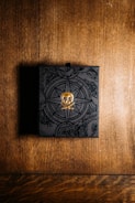 A black box with intricate designs and a golden skull emblem on top sits on a wooden surface. The background features a warm, textured wood pattern, and the box appears to have a mysterious, ornate design, possibly indicating a theme of adventure or exploration.