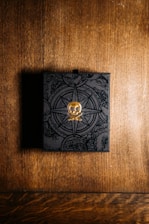 A black box with intricate designs and a golden skull emblem on top sits on a wooden surface. The background features a warm, textured wood pattern, and the box appears to have a mysterious, ornate design, possibly indicating a theme of adventure or exploration.