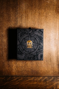 A black box with intricate designs and a golden skull emblem on top sits on a wooden surface. The background features a warm, textured wood pattern, and the box appears to have a mysterious, ornate design, possibly indicating a theme of adventure or exploration.