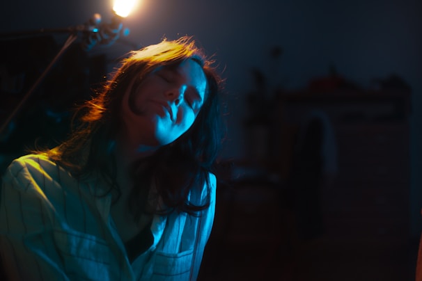 A person with long hair is sitting with their eyes closed, illuminated by soft, warm lighting from a lamp overhead. They are wearing a striped shirt, and their expression appears serene and relaxed. The setting is dimly lit with visible elements of a room in the background.