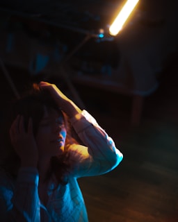 A woman dressed in a striped shirt touches her head with both hands, illuminated by dramatic lighting that casts deep shadows and highlights. The surroundings appear dim, with a warm, bright light source above her adding a moody atmosphere.