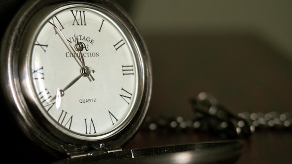 A vintage pocket watch is displayed with a silver casing and Roman numeral markings on the face. The words 'Vintage Collection' and 'Quartz' are visible. The background is softly blurred, focusing attention on the watch.
