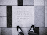 The image depicts a close-up view of a tiled sidewalk where a person wearing black shoes is standing. The tiles are arranged in a pattern with alternating black and white colors. One of the tiles has an engraved text: 'FOR GOD HAS NOT GIVEN US A SPIRIT OF FEAR AND TIMIDITY, BUT OF POWER, LOVE, AND SELF-DISCIPLINE. 2 TIMOTHY 1:7'.