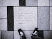 The image depicts a close-up view of a tiled sidewalk where a person wearing black shoes is standing. The tiles are arranged in a pattern with alternating black and white colors. One of the tiles has an engraved text: 'FOR GOD HAS NOT GIVEN US A SPIRIT OF FEAR AND TIMIDITY, BUT OF POWER, LOVE, AND SELF-DISCIPLINE. 2 TIMOTHY 1:7'.