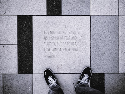 The image depicts a close-up view of a tiled sidewalk where a person wearing black shoes is standing. The tiles are arranged in a pattern with alternating black and white colors. One of the tiles has an engraved text: 'FOR GOD HAS NOT GIVEN US A SPIRIT OF FEAR AND TIMIDITY, BUT OF POWER, LOVE, AND SELF-DISCIPLINE. 2 TIMOTHY 1:7'.