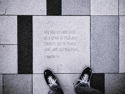 The image depicts a close-up view of a tiled sidewalk where a person wearing black shoes is standing. The tiles are arranged in a pattern with alternating black and white colors. One of the tiles has an engraved text: 'FOR GOD HAS NOT GIVEN US A SPIRIT OF FEAR AND TIMIDITY, BUT OF POWER, LOVE, AND SELF-DISCIPLINE. 2 TIMOTHY 1:7'.