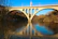 A sturdy, newly constructed bridge arching over a calm river with workers inspecting the structure.