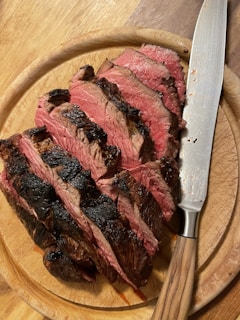 Several slices of cooked meat are displayed on a round wooden cutting board. The meat appears to be juicy with a charred crust and is cooked to a medium-rare level of doneness. A knife with a wooden handle rests beside the slices.