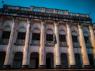 An old colonial-style building with a series of tall, ornate columns. The facade displays intricate detailing and weathered textures, suggesting historical significance. A flag stands in front of the structure.