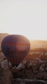 A hot air balloon is seen floating over a rugged landscape during sunset or sunrise, casting a warm glow over the scene. The balloon has 'Sultan' written on it and is prominent against the backdrop of rocky terrain and a clear sky.