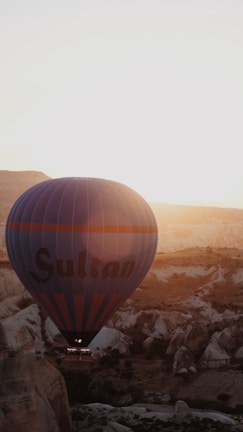 A hot air balloon is seen floating over a rugged landscape during sunset or sunrise, casting a warm glow over the scene. The balloon has 'Sultan' written on it and is prominent against the backdrop of rocky terrain and a clear sky.