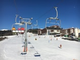 Ski resort scene with a chairlift carrying several skiers in colorful jackets. Snow-covered slopes and buildings in the background, with skiers enjoying the slopes. Clear blue skies suggest a sunny day.
