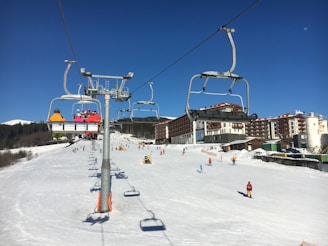 Ski resort scene with a chairlift carrying several skiers in colorful jackets. Snow-covered slopes and buildings in the background, with skiers enjoying the slopes. Clear blue skies suggest a sunny day.