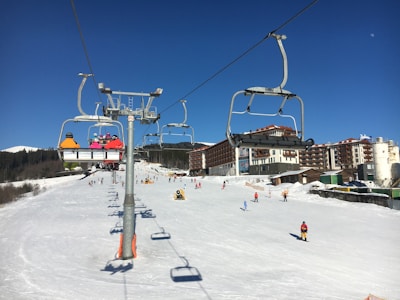 Ski resort scene with a chairlift carrying several skiers in colorful jackets. Snow-covered slopes and buildings in the background, with skiers enjoying the slopes. Clear blue skies suggest a sunny day.