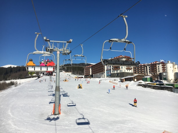 Ski resort scene with a chairlift carrying several skiers in colorful jackets. Snow-covered slopes and buildings in the background, with skiers enjoying the slopes. Clear blue skies suggest a sunny day.
