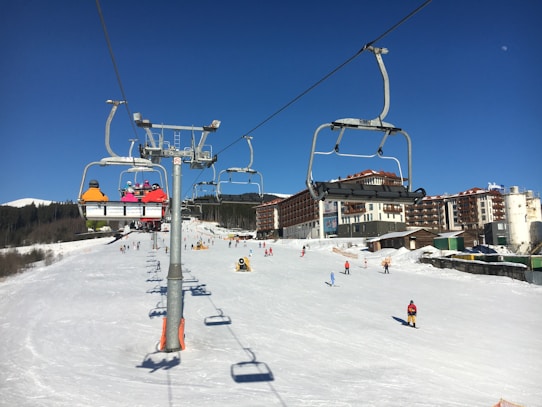 Ski resort scene with a chairlift carrying several skiers in colorful jackets. Snow-covered slopes and buildings in the background, with skiers enjoying the slopes. Clear blue skies suggest a sunny day.