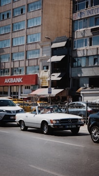 A white convertible vintage Mercedes-Benz is parked on a city street alongside other modern vehicles. The backdrop consists of a row of mid-rise buildings with a visible sign for Akbank. The scene conveys a juxtaposition of classic and modern elements within an urban environment.