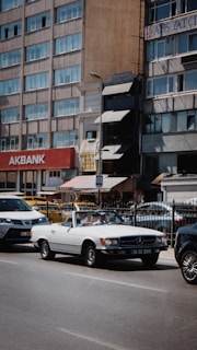 A white convertible vintage Mercedes-Benz is parked on a city street alongside other modern vehicles. The backdrop consists of a row of mid-rise buildings with a visible sign for Akbank. The scene conveys a juxtaposition of classic and modern elements within an urban environment.
