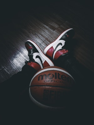 A pair of stylish red, white, and black sneakers rest on a dark wooden floor. In front of them lies a Molten official basketball with FIBA branding. The lighting is dim, creating a dramatic contrast between the objects and their surroundings.