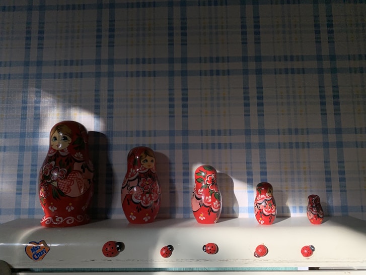 A set of Russian nesting dolls is arranged in a line on a shelf. Each doll features vibrant red and black floral patterns with painted faces. The background is a blue and white checkered wallpaper, and the shelf is adorned with small ladybug decorations.