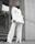 A person stands confidently wearing a stylish white suit and a black scarf. The background features concrete walls and green plants, creating a modern urban setting. Their stance and expression convey a sense of empowerment and fashion-forward style.