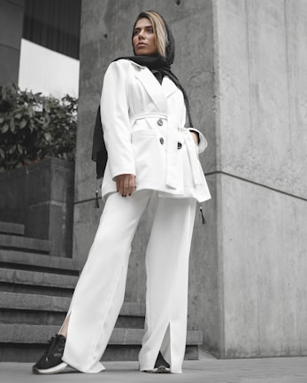 A person stands confidently wearing a stylish white suit and a black scarf. The background features concrete walls and green plants, creating a modern urban setting. Their stance and expression convey a sense of empowerment and fashion-forward style.