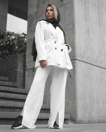 A person stands confidently wearing a stylish white suit and a black scarf. The background features concrete walls and green plants, creating a modern urban setting. Their stance and expression convey a sense of empowerment and fashion-forward style.