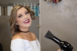 A smiling person with wavy hair holds a black hair dryer in a salon setting. The background shows shelves with various bottles of hair products.