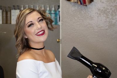 A smiling person with wavy hair holds a black hair dryer in a salon setting. The background shows shelves with various bottles of hair products.