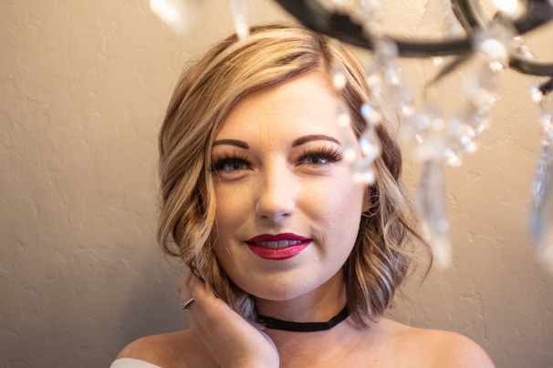 A person with short blonde hair and red lipstick stands in front of a neutral background. Crystal decorations hang in front, partially out of focus, adding an elegant touch. The person's expression is calm and their hand gently touches their face.