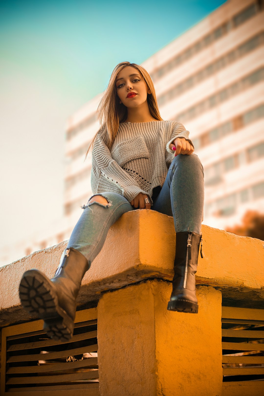 woman in gray sweater and blue denim jeans sitting on brown concrete wall during daytime