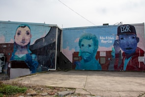 The image showcases a large mural painted on the side of a building featuring three distinct figures. The mural on the left portrays a stylized portrait of a person with large eyes and a blue hood. In the middle, there is an abstract depiction of a man with a beard, rendered in a blue-green color. On the right, a figure wearing a cap with the text 'D2R' is shown in a vibrant mix of colors including red and blue. Graffiti-style text accompanies the mural.