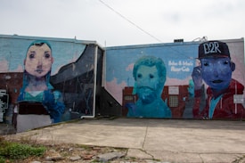 The image showcases a large mural painted on the side of a building featuring three distinct figures. The mural on the left portrays a stylized portrait of a person with large eyes and a blue hood. In the middle, there is an abstract depiction of a man with a beard, rendered in a blue-green color. On the right, a figure wearing a cap with the text 'D2R' is shown in a vibrant mix of colors including red and blue. Graffiti-style text accompanies the mural.