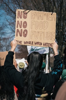 A person is holding up a cardboard protest sign that reads 'NO NO NO NO FARMER FOOD FUTURE THE WHOLE WORLD WATCHES'. The person has long dark hair and is wearing a black hoodie. Other protesters are visible in the background holding similar signs, suggesting a rally or demonstration. Leafless trees are visible, indicating it might be early spring or late fall.