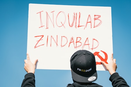 A person is holding a sign with red handwritten text 'INQUILAB ZINDABAD' and a red communist symbol on a white background against a clear blue sky. The individual is wearing a black cap with the word 'FREEDOM' in white letters.