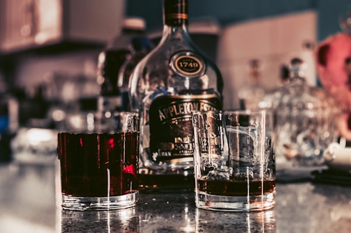 Two glasses with drinks are placed in front of a bottle of alcohol on a reflective countertop. The bottle is prominent with a dark label, and the background is softly blurred with various bottles and kitchen elements visible.
