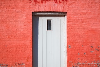 A wooden door with vertical panels set into a vivid red brick wall. The door is painted white and features a small rectangular window at the top. There are some signs of wear and tear on the wall, with spots where the paint has chipped away.